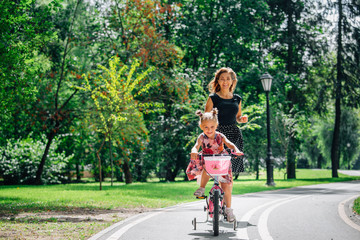 Beautiful and happy young mother teaching her cute daughter to ride a bicycle. Both smiling and looking at each other. Summer park in background. Happy parenting concept.