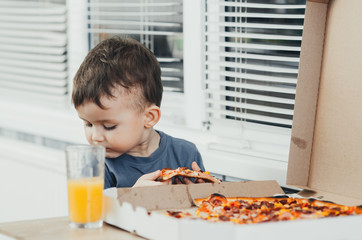 the little boy eats a huge harmful pizza himself in the kitchen and drinks juice, very fat and harmful