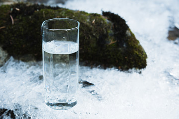 Transparent glass glass with drinking mountain water in winter stands on an icy crust against the background of a clean, frosty river bank. The concept of drinking mountain drinking mineral water and