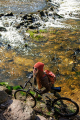 long haired redhead young woman in red cap riding a bike near mountain streamy river in spring