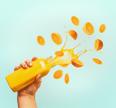 Female Hand Holding Bottle With Yellow Splash Summer Beverage: Smoothie Or Juice And Flying Oranges At Blue Background.