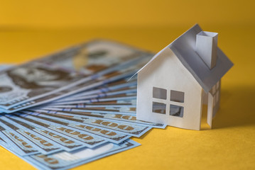Small paper house on pile of dollar bills on yellow background. Real estate prices concept