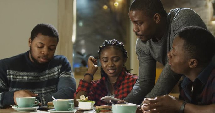 Group of laughing African-American men and girl watching phone and laughing while chilling in cafe.