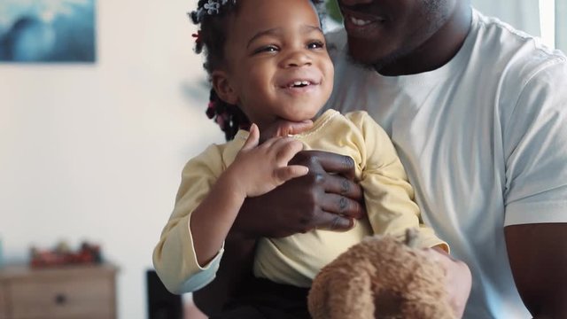 Sweet little African American baby-girl is held on her father’s arms, laughs and tries to get away from his hugs. Favorite toy, positive emotions. Slow motion, close up view