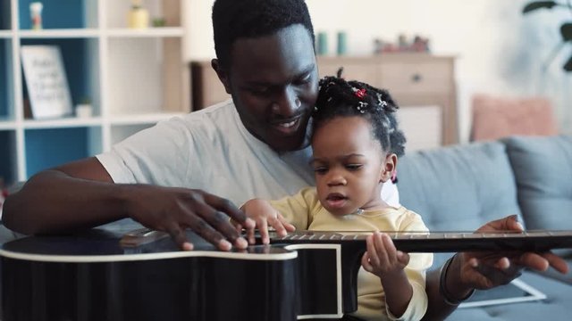 Smiling young father holds his little cute baby-girl on his knees and tries to teach her to play the guitar, cheerful toddler claps the guitar as if she playing it. Modern family, happy memories. Slow