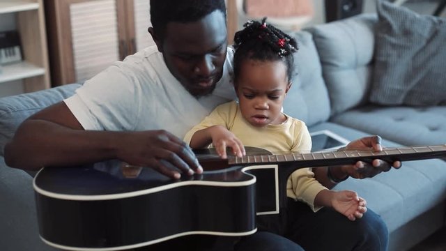 Close up view of adorable little baby-girl sitting on her father’s knees and actively stroking the guitar, as if she plays it. Positive emotions, family bonds. Cozy living room. Slow motion