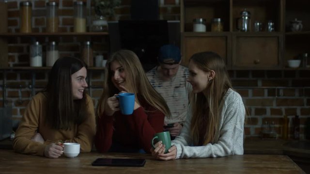 Cheerful young female friends discussing latest gossips over coffee while relaxing in domestic kitchen. Three beautiful young women talking and smiling while drinking coffee in the kitchen.