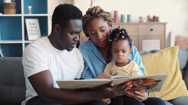Cute young parents turn the pages of the book for their daughter, little adorable baby attentively looks at the colorful pictures, they are discussing it. Family time, family portrait. Close up view