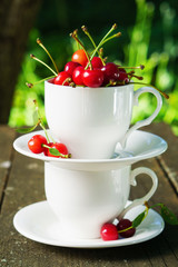 Cherries with leaves in the beautiful white cup on brown wooden rough rural table. Summer rustic garden view. Vertical, selective focus.