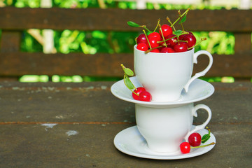 Cherries with leaves in the beautiful white cup on brown wooden rough rural table. Summer rustic garden view. Copy space, selective focus.