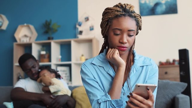 Young woman uses her smartphone, posts photos, reads blogs, while her husband on the background tries to keep calm their daughter, woman gets immersed in thought. Close up view