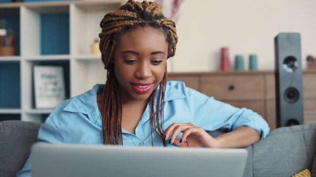 Attractive African American young woman with stylish dreadlocks actively uses the laptop, surfs the internet, smiles towards the screen. Being online, blogger, modern communication. Close up view