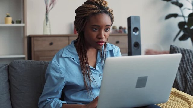 Beautiful African American young woman with stylish dreadlocks sits on the couch in light living room and actively uses the laptop, types the messages. Relaxation, blogger, modern communication. Close
