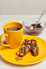 Top view of  three, freshly baked, homemade, peanut butter oatmeal biscotti on a round, yellow plate with a large yellow cup of hot, coffee and a glass bowl of chocolate frosting