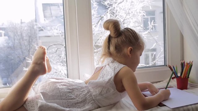 Little girl draws pictures lying on the window sill. Sunny winter morning