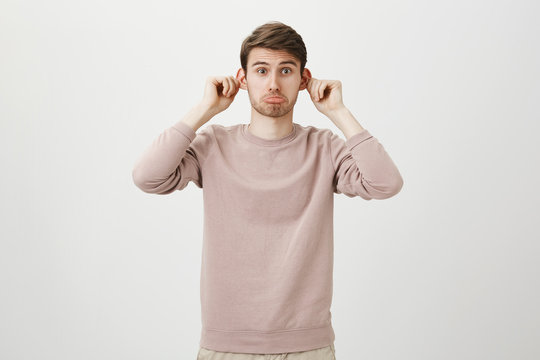 Indoor portrait of attractive funny european male stretching ears and sticking out tongue, imitating monkey and looking at camera while standing over gray background. Guy looks in mirror to cheer up