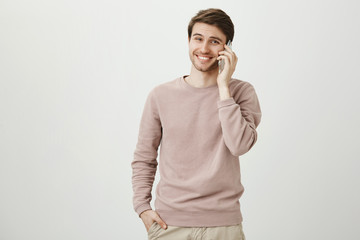 Positive and handsome young caucasian man with bristle smiling while talking on smartphone, standing with hand in pocket over gray background. Guy talks with his little sister who misses him