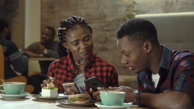 Attractive young black couple in a cafeteria enjoying cups of coffee together and browsing social media on their mobile phone in a close up view.