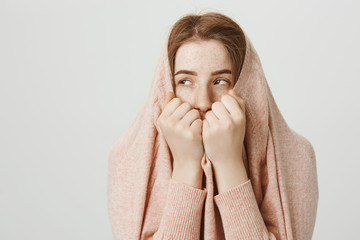 Portrait of bothered or annoyed teenage ginger girl hiding in pullover while sticking face out of collar, looking aside with bored expression against gray background. Woman escapes from stares