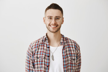 Charming shop assistant ready to offer his help. Portrait of attractive emotive young man with beard in eyewear smiling cheerfully while posing against gray background in studio. Emotions concept