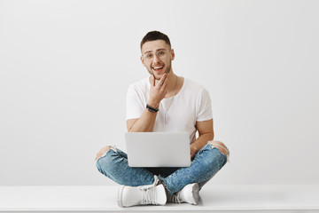 Should I write this post in my blog or not. Studio portrait of emotive handsome man with bristle and eyewear sitting with crossed legs on floor holding notebook on laps, touching chin while thinking