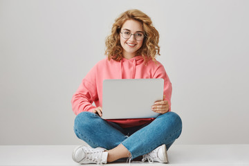 Help me with homework. Portrait of happy interesting girl with curly hair, working on project, sitting on floor with crossed legs, holding laptop and smiling broadly, freelancing over gray background