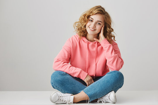 Studio portrait of cute female freelancer with curly hair sitting on floor with crossed lefs leaning head on hand and smiling at camera. Singer with band discuss new song they want to release