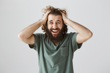 Bad things happen to good people. Portrait of stunned and stressed mature hispanic guy with curly hair and beard shouting from pain or depression, holding hands on head, being miserable over gray wall