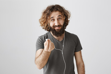 Find someone who will share earphones with you. Portrait of handsome friendly eastern guy with curly hair pulling earbud at camera, wanting to listen music together with mate while in subway