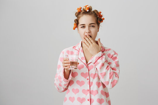 Exhausted cute european wife wearing hair curlers and pajamas with heart template, yawning and covering mouth with hand, drinking glass of water over gray background. Tired sister heading to her room