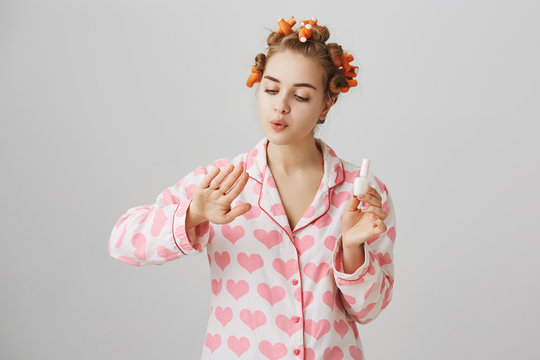 Glamourous girl at home wearing hair curlers and lovely pajamas in hearts, blowing at fingers while painting nails, being focused over gray background. Feminine girlfriend looks after her appearance