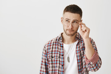 Guy trying to make calculations in mind. Indoor shot of good-looking modern male with beard, wearing and holding eyewear while looking up with thoughtful expression, making decision over gray wall