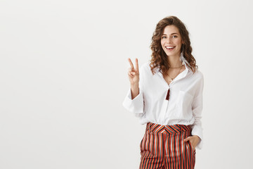 Portrait of confident successful woman in stylish garment showing v-sign or peace gesture while smiling broadly and expressing satisfaction with result, standing over gray background.
