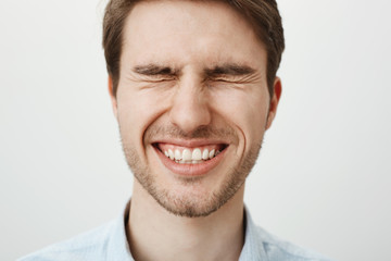 Emotive happy european guy with bristle standing with tightly closed eyes, squinting and smiling broadly, waiting for surprise or making wish near birthday cake, standing over gray background