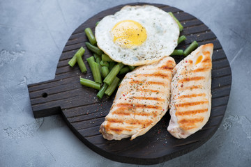 Black wooden serving board with grilled chicken breasts, fried egg and green beans, studio shot on a grey concrete background, selective focus