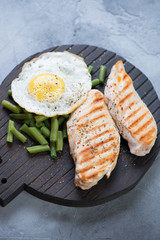 Grilled chicken breast fillet with green beans and fried egg on a black wooden chopping board, selective focus, vertical shot, elevated view