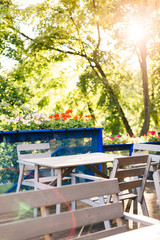 Wooden table and chairs on cafe terrace. Open restaurant terrace with flowers under big green trees. Summer, outdoors.