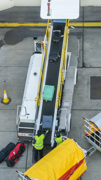 High Perspective Of Unidentifitable Baggage Handlers Picking Up Suitcases From A Conveyor Belt Connected To A Commercial Airplane.