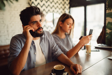 A beautiful young woman is upset to her handsome boyfriend because he is talking on the phone while they are having a coffee on the table.