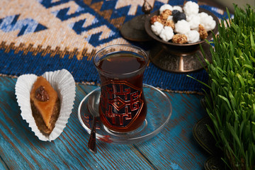 Pakhlava or baklava with glass of black tea and sugar sweet snacks in silver bowl on wooden rustic table with wheat grass semeni and blue carpet. Novruz holiday 