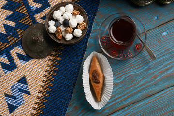 Pakhlava or baklava with glass of black tea and sugar sweet snacks in silver bowl on wooden rustic table with wheat grass semeni and blue carpet. Novruz holiday 