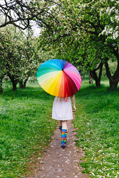 Girl walking under the big colorful rainbow-umbrella in the blooming garden. Spring, outdoors.