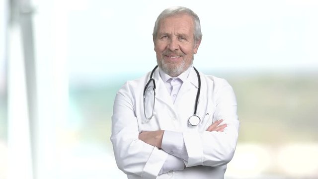 Happy senior doctor on blurred background. Happy male elderly doctor with crossed arms. People, professions and healthcare.