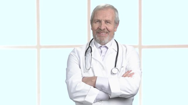 Male bearded doctor on blurred background. Senior european doctor in white coat folded arms on window background. Happy mature doctor, portrait.