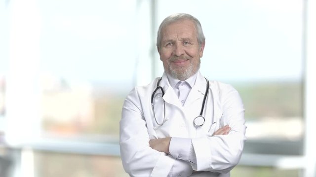 Elderly doctor with stethoscope. Happy senior cardiologist folded arms on blurred background. People, profession, medicine.