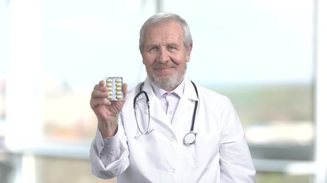 Senior doctor in white uniform showing pills. Mature doctor with blister of tablets showing thumb up, blurred background. Profession, people and healthcare.