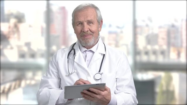 Senior smiling doctor with pc tablet. Elderly bearded doctor working on his tablet at work. People, technology, connection.