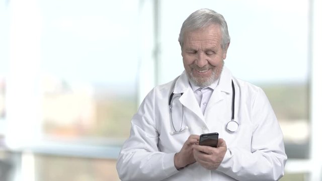 Smiling doctor using his smartphone. Joyful elderly doctor typing a message on his smartphone on abstract blurred background.
