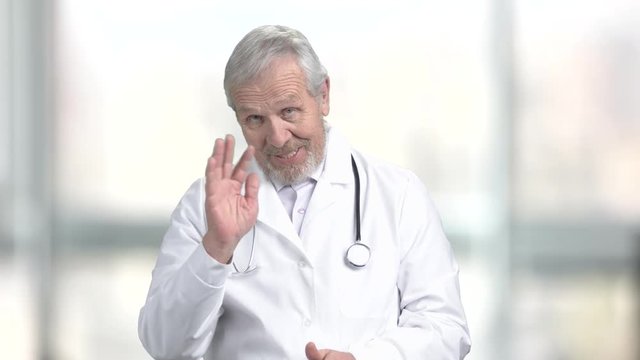 Cheerful senior doctor talking to camera. Happy medical worker waving with hand and looking at camera, blurred background.