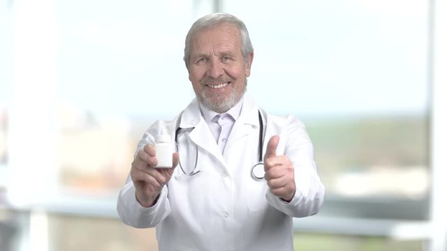 Senior doctor holding bottle of pills. Smiling elderly doctor with pills showing thumb up on blurred background. Symbol of success.